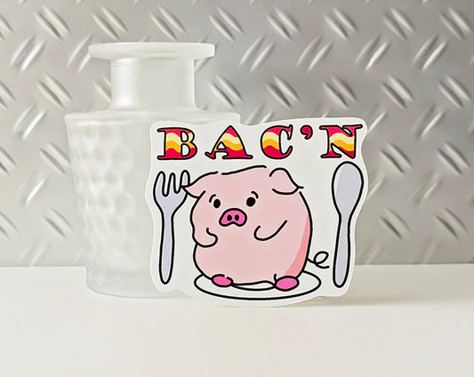 Sticker with a cartoon pig and 'BAC'N' text on a white surface with a metallic background